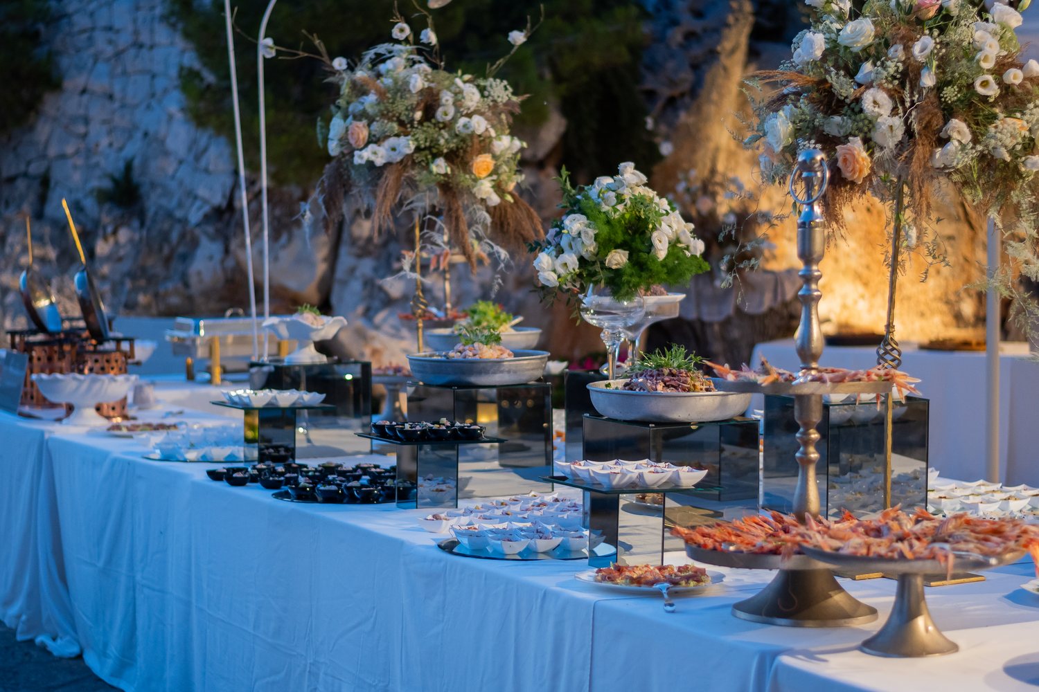 Il buffet di crudi di mare e antipasti allestito davanti alla parete di roccia, tra alte composizioni floreali — Cala dei Balcani, Santa Cesarea Terme, Salento