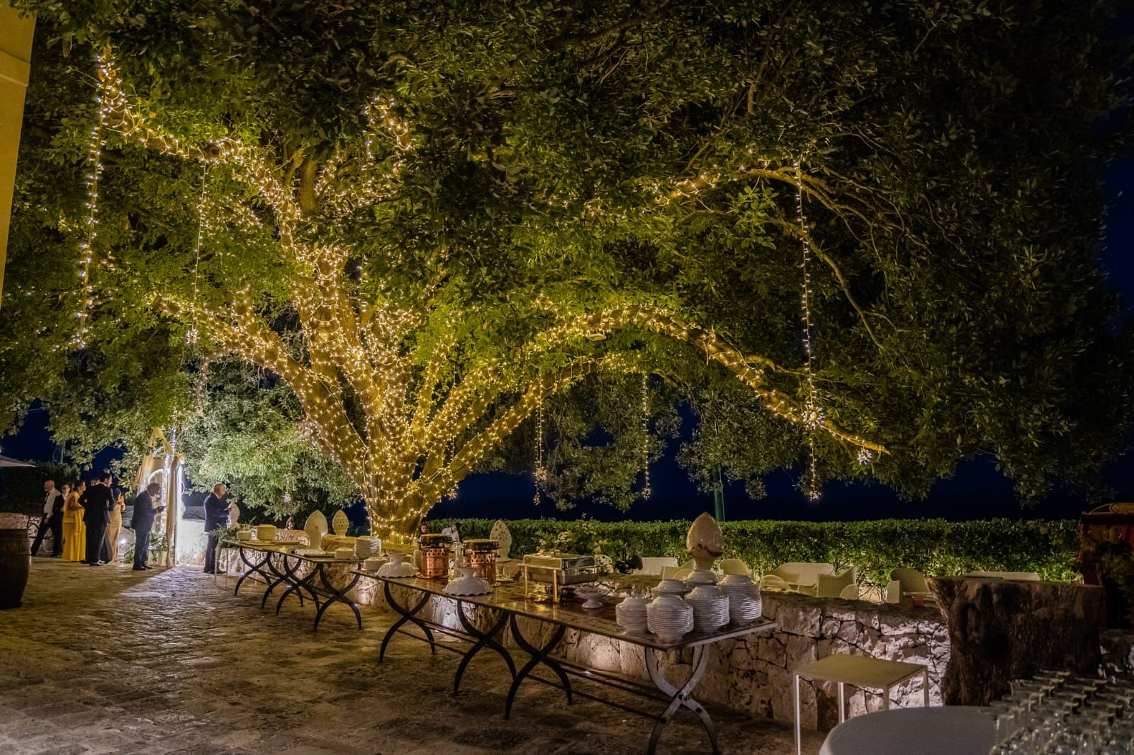 Il Giardino della Quercia illuminato di sera con i tavoli del buffet del ricevimento — Cala dei Balcani, Santa Cesarea Terme, Salento