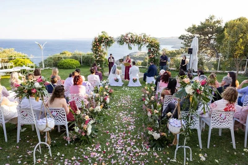 Cerimonia all'aperto con arco di fiori rosa e petali sul prato — destination wedding internazionale a Cala dei Balcani, Salento