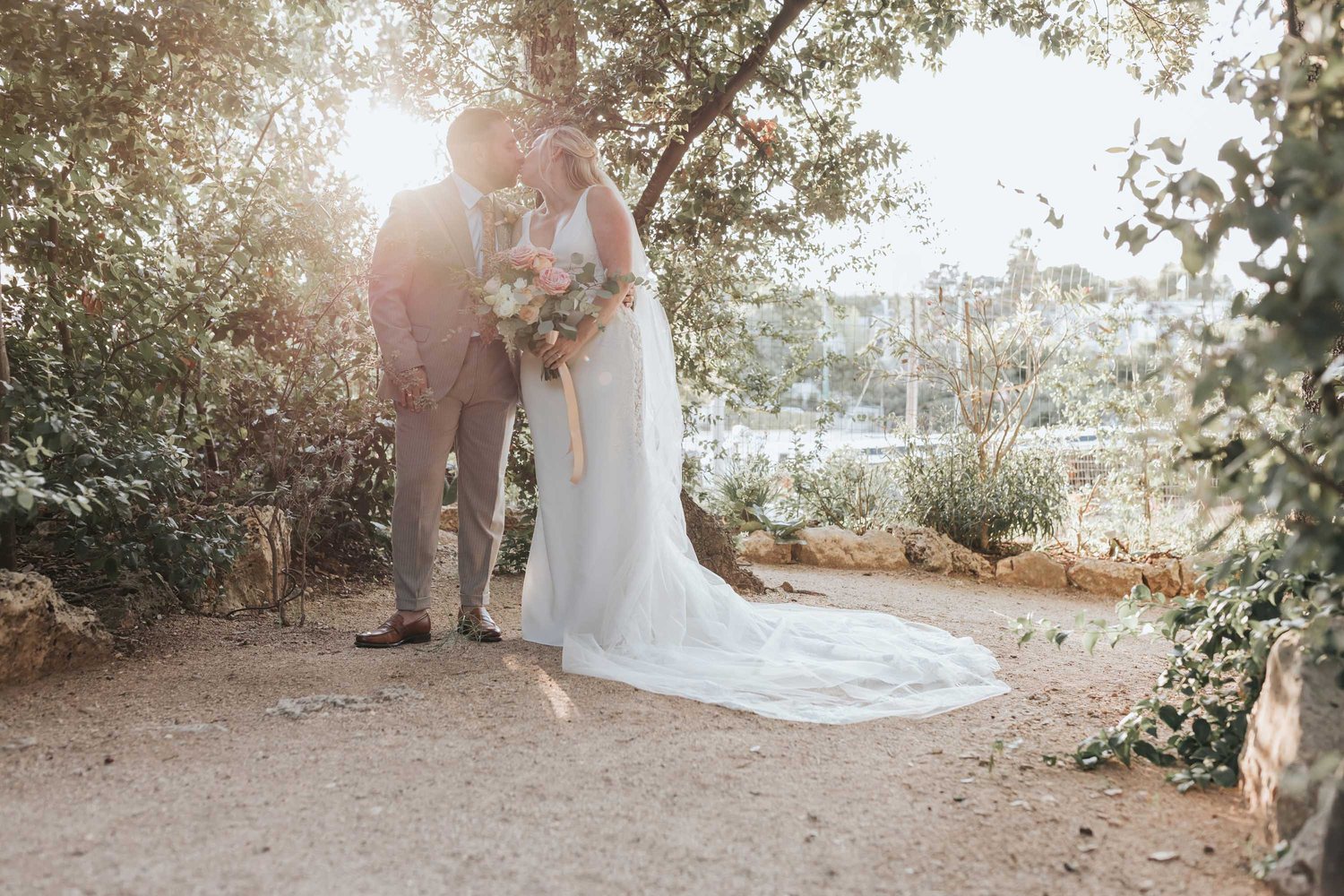 Les mariés s&rsquo;embrassent à contre-jour dans le jardin, la longue traîne sur l&rsquo;allée de pierre &mdash; Cala dei Balcani, Santa Cesarea Terme, Salento