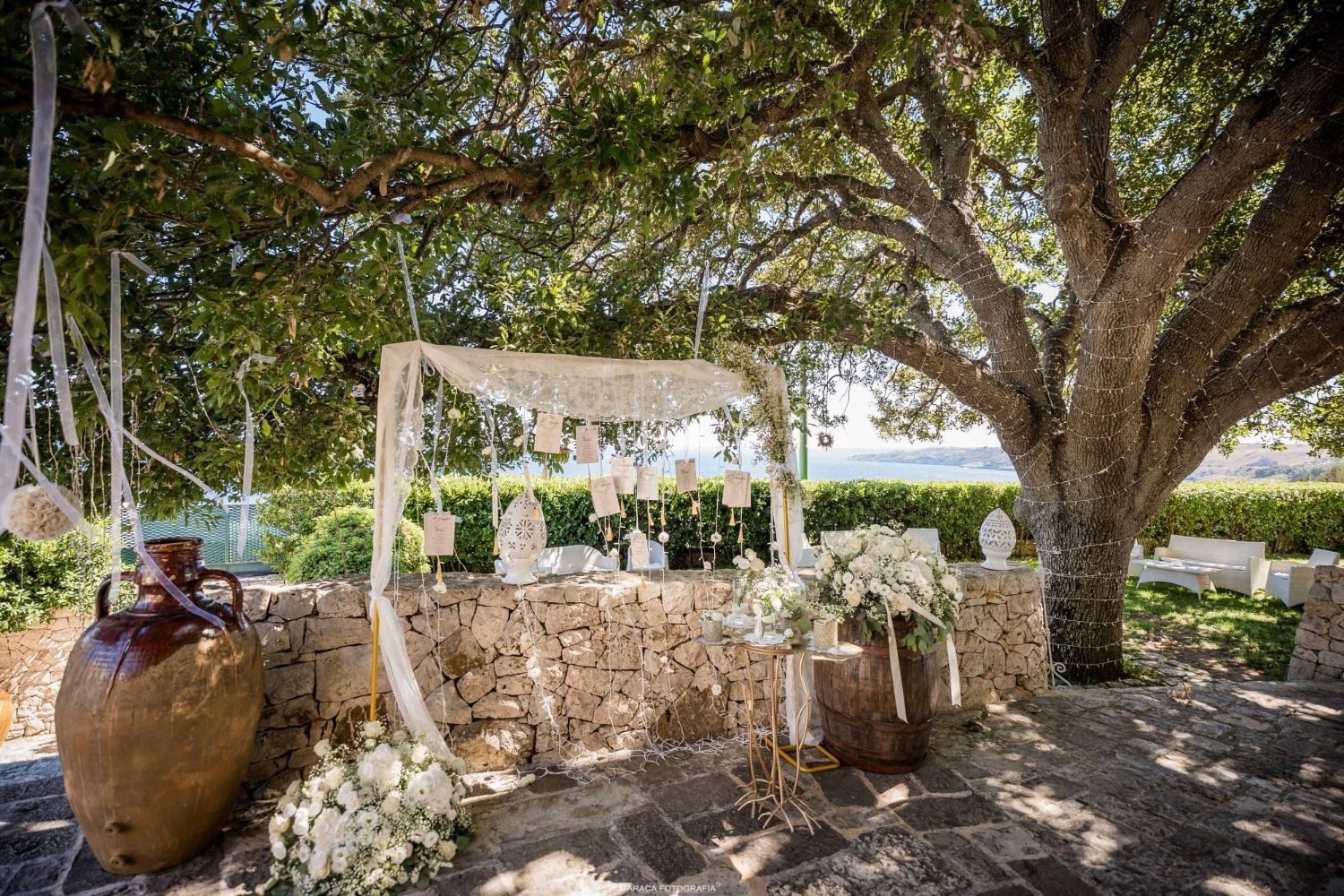 Tableau de mariage sotto l'albero secolare, con drappo di pizzo e anfora in terracotta, vista sulla baia — Cala dei Balcani, Salento