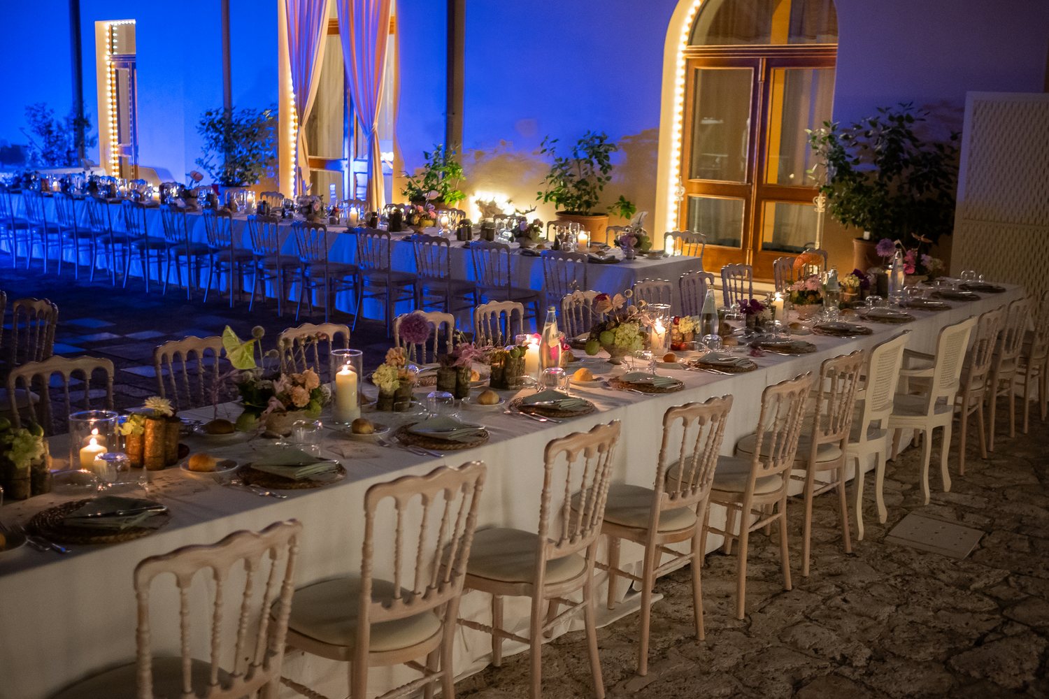 Tavolata imperiale allestita al calar della sera, illuminata di azzurro tra candele e composizioni basse — Cala dei Balcani, Santa Cesarea Terme, Salento