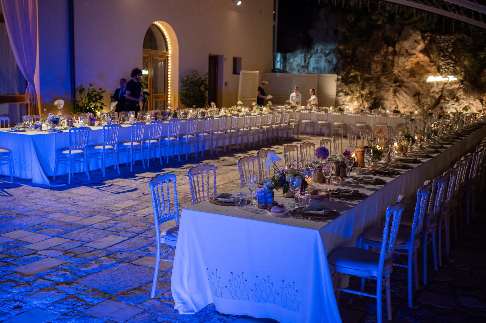 Tavolata imperiale allestita di sera nel cortile, illuminata da luci blu — ricevimento di matrimonio a Cala dei Balcani, Salento