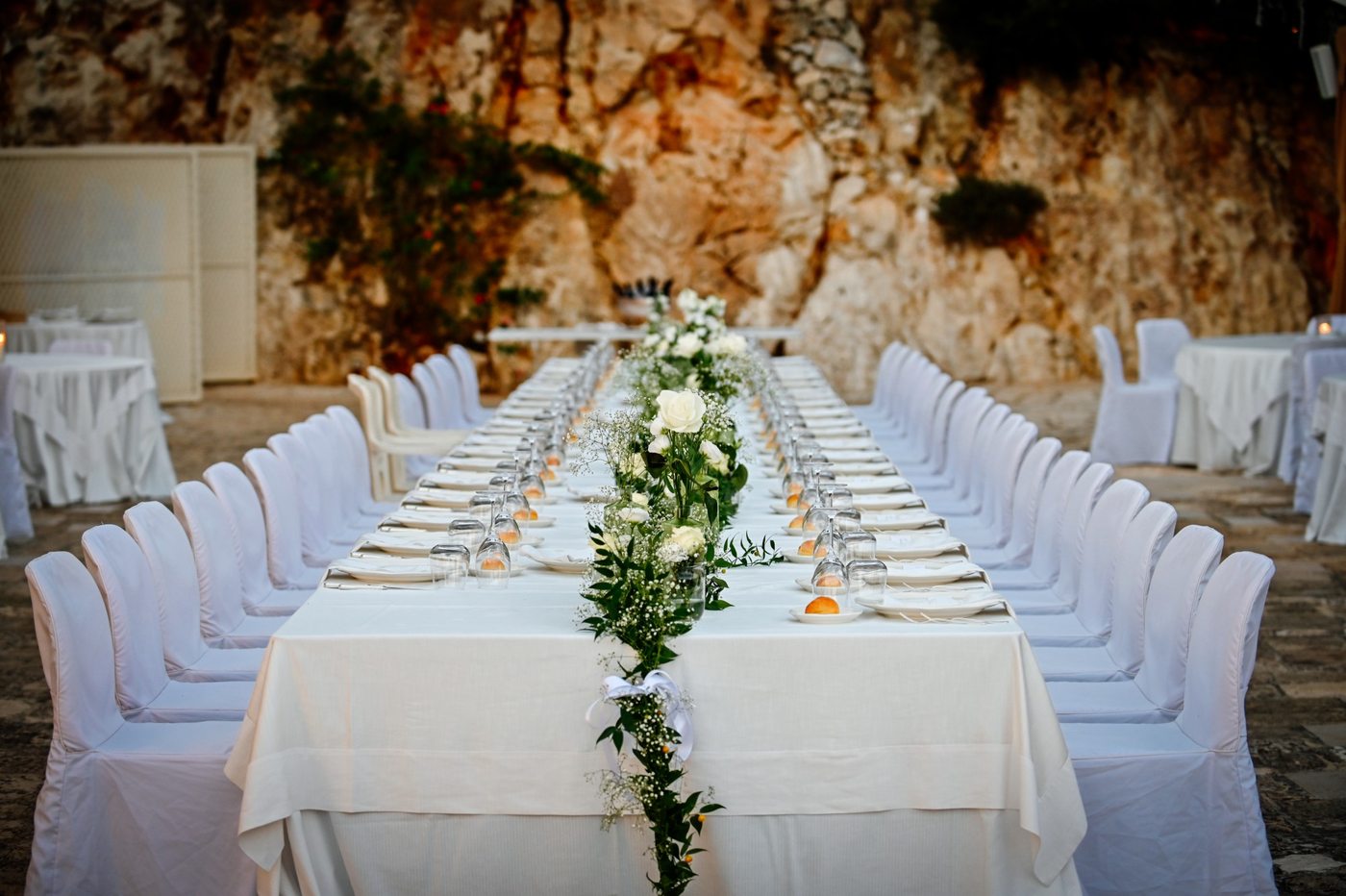 Une seule longue table pour un mariage intime devant la paroi rocheuse, avec un chemin de roses blanches et de gypsophile &mdash; Cala dei Balcani, Salento