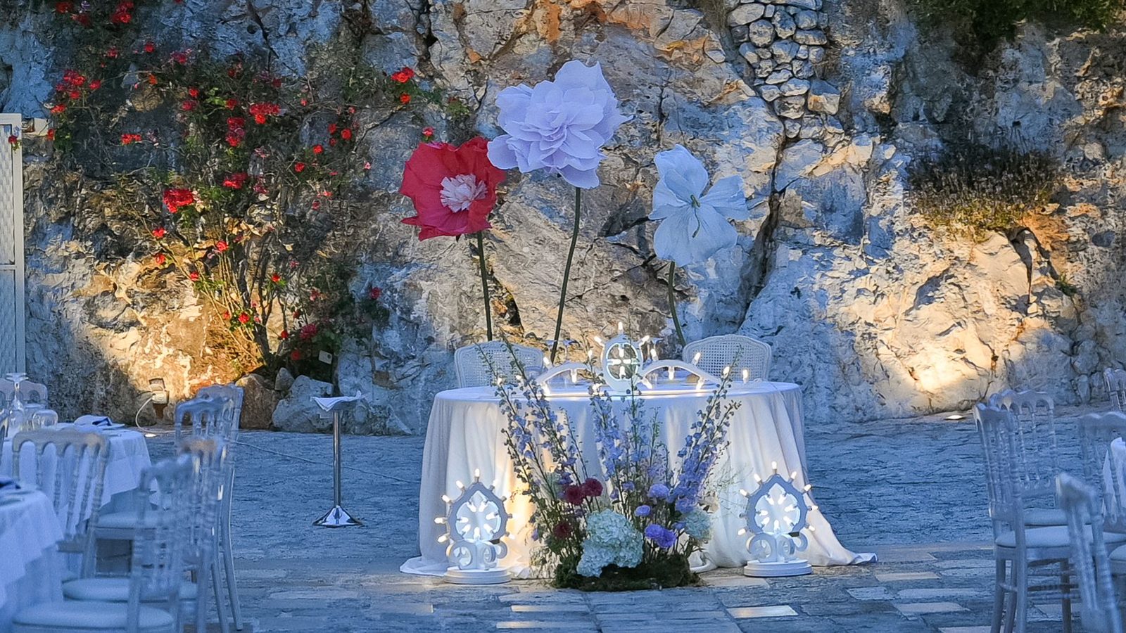 Tavolo degli sposi illuminato davanti alla parete rocciosa, con grandi fiori di carta e composizioni di campo — Cala dei Balcani, Salento